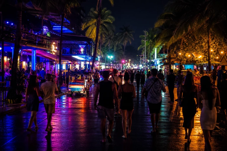 Rue animée de Phuket la nuit avec néons, palmiers et voyageurs devant des clubs, sans texte visible.