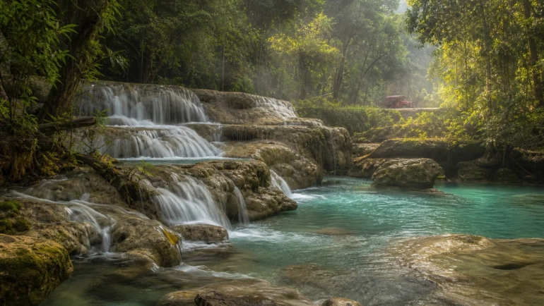 A lush waterfall near Chiang Mai surrounded by tropical forest and limestone rocks, with a red songthaew visible in the dista